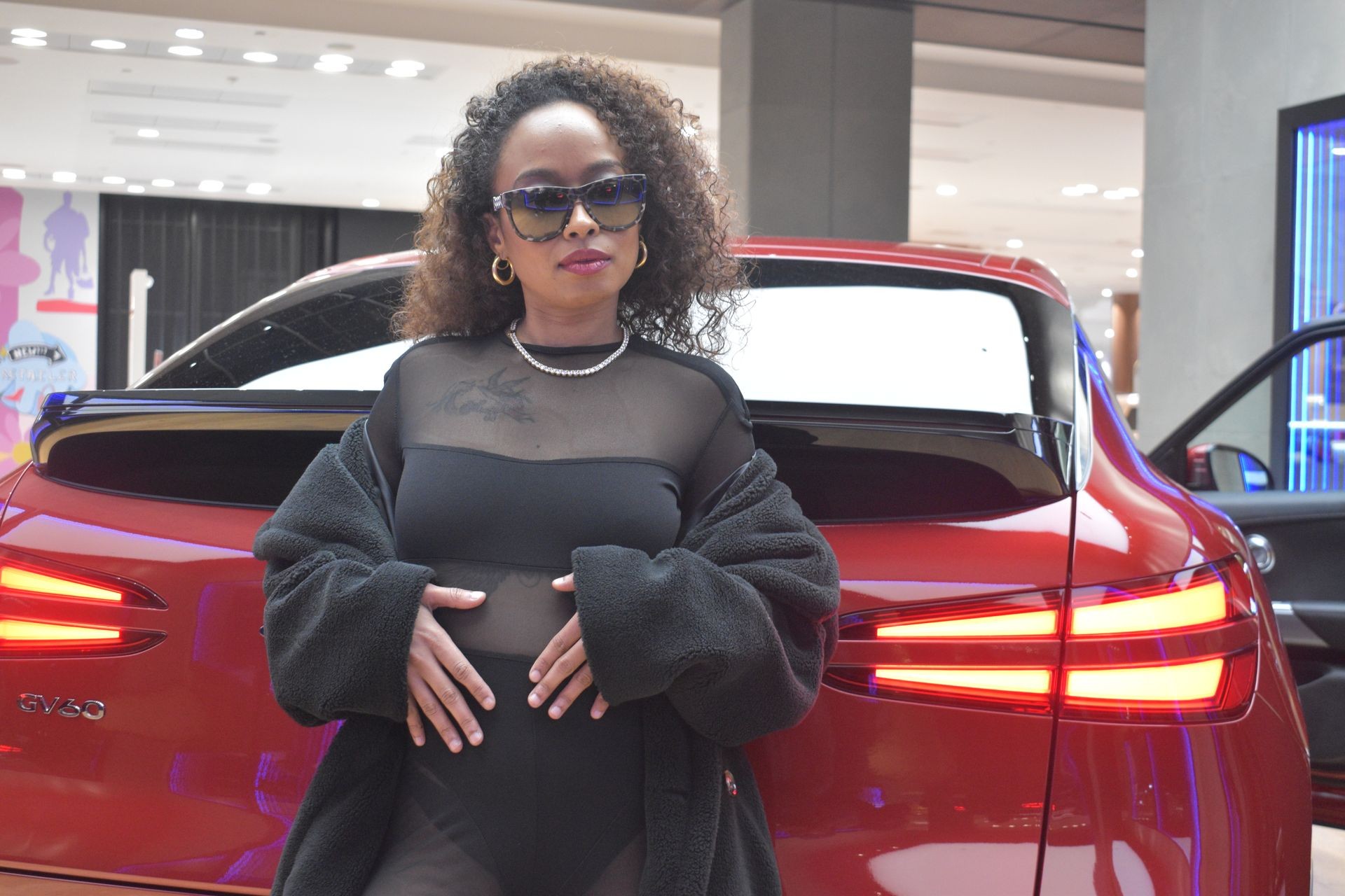 Person wearing sunglasses posing in front of a red car indoors.