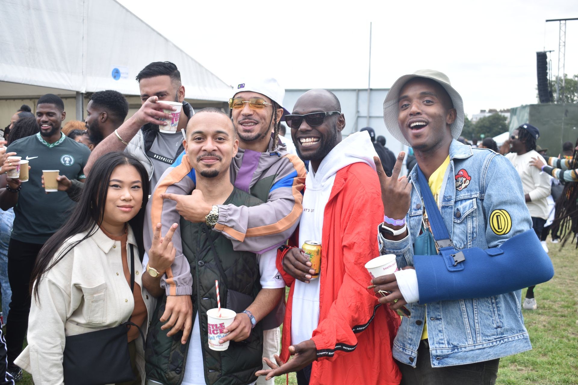 Group of friends smiling and holding drinks at an outdoor event, one person with arm in sling.