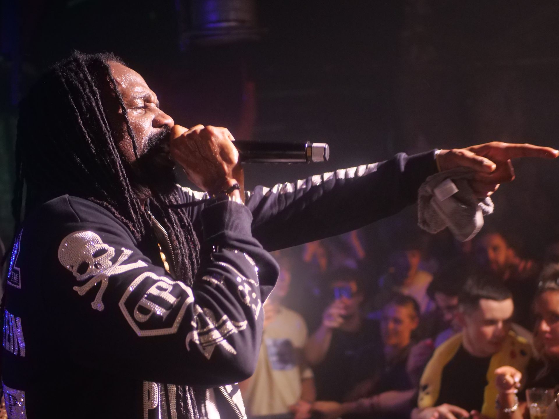 Performer with dreadlocks and skull-patterned jacket singing on stage, pointing towards the audience.