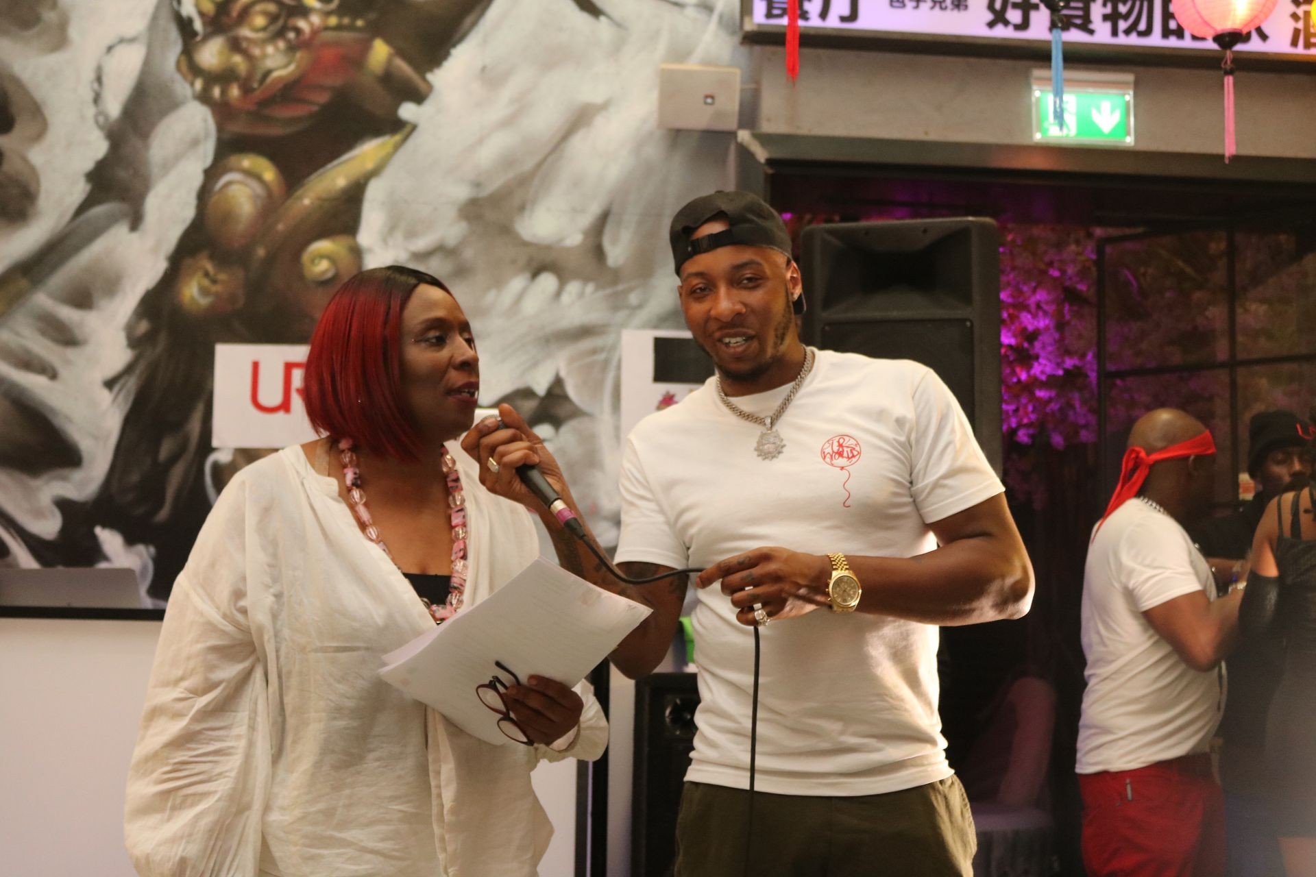 Two people speaking into microphones at an indoor event, with a colorful mural in the background.