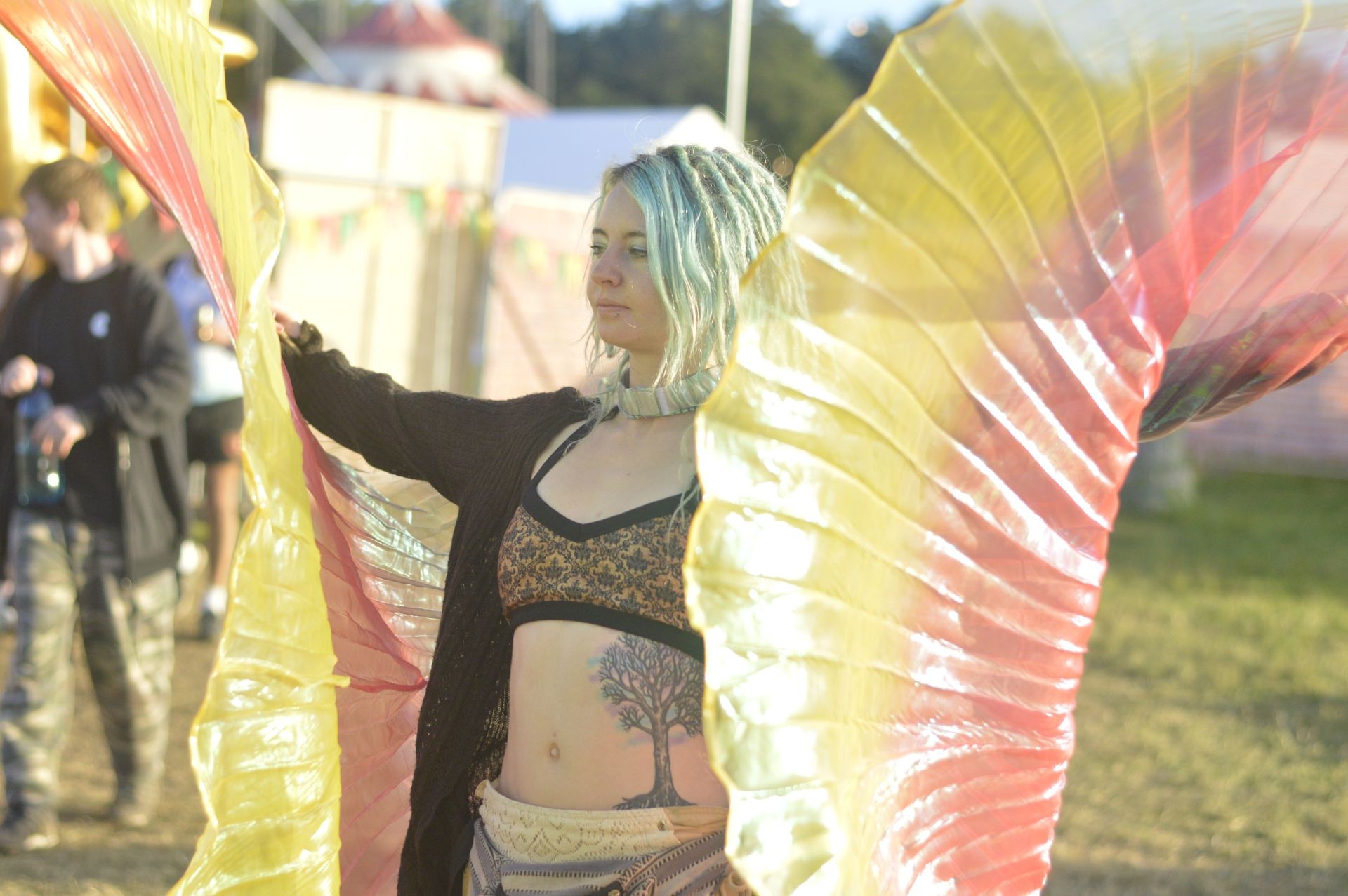 Person with colorful wings dancing at an outdoor festival, wearing a patterned top and dreadlocks hairstyle.
