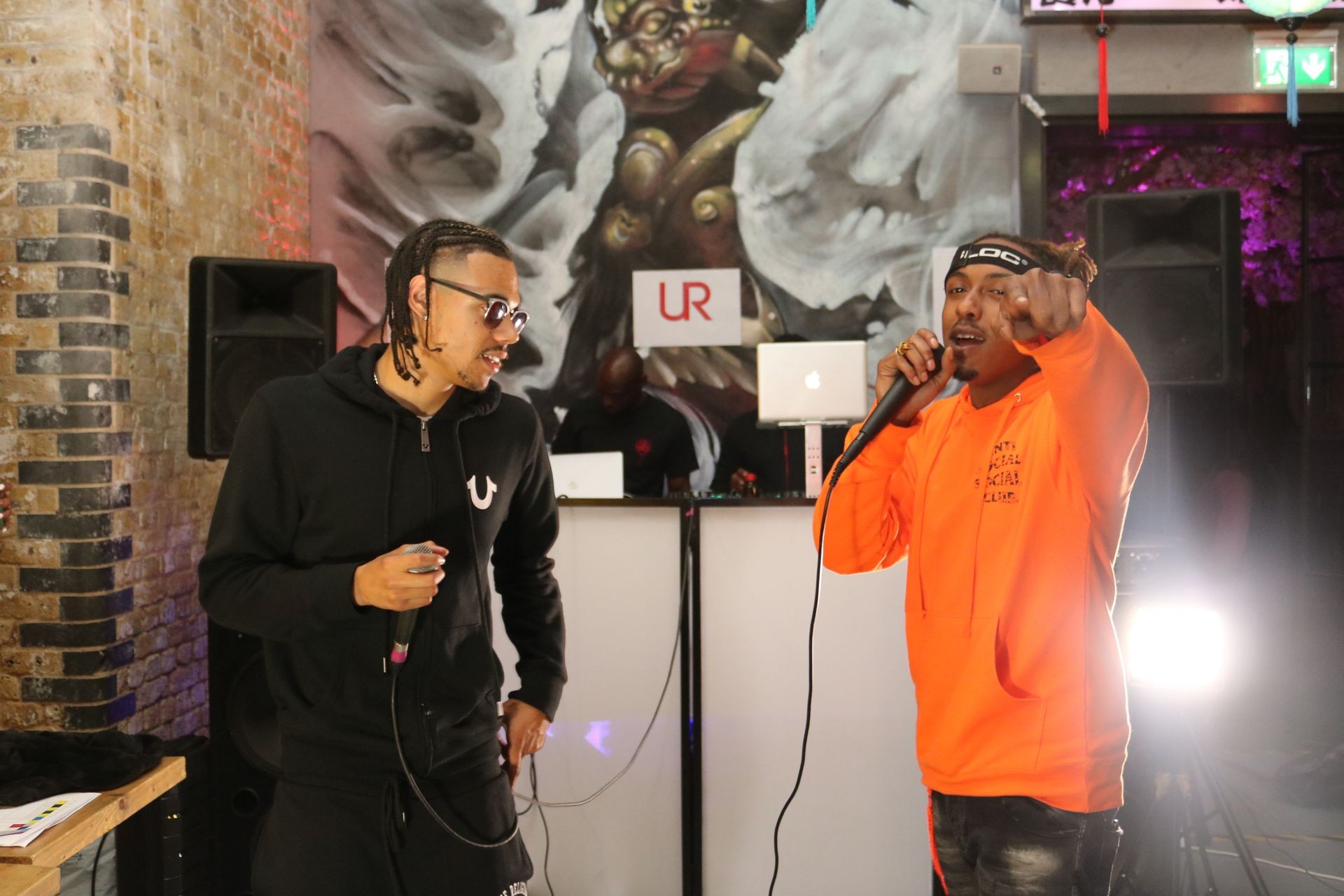 Two performers holding microphones on stage with a colorful mural and sound equipment in the background.